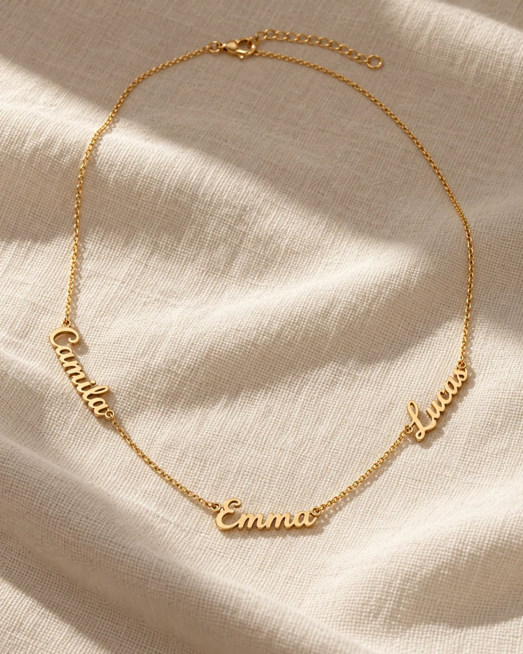Gold name necklace with three cursive names, Camila, Emma and Lucas, on linen
