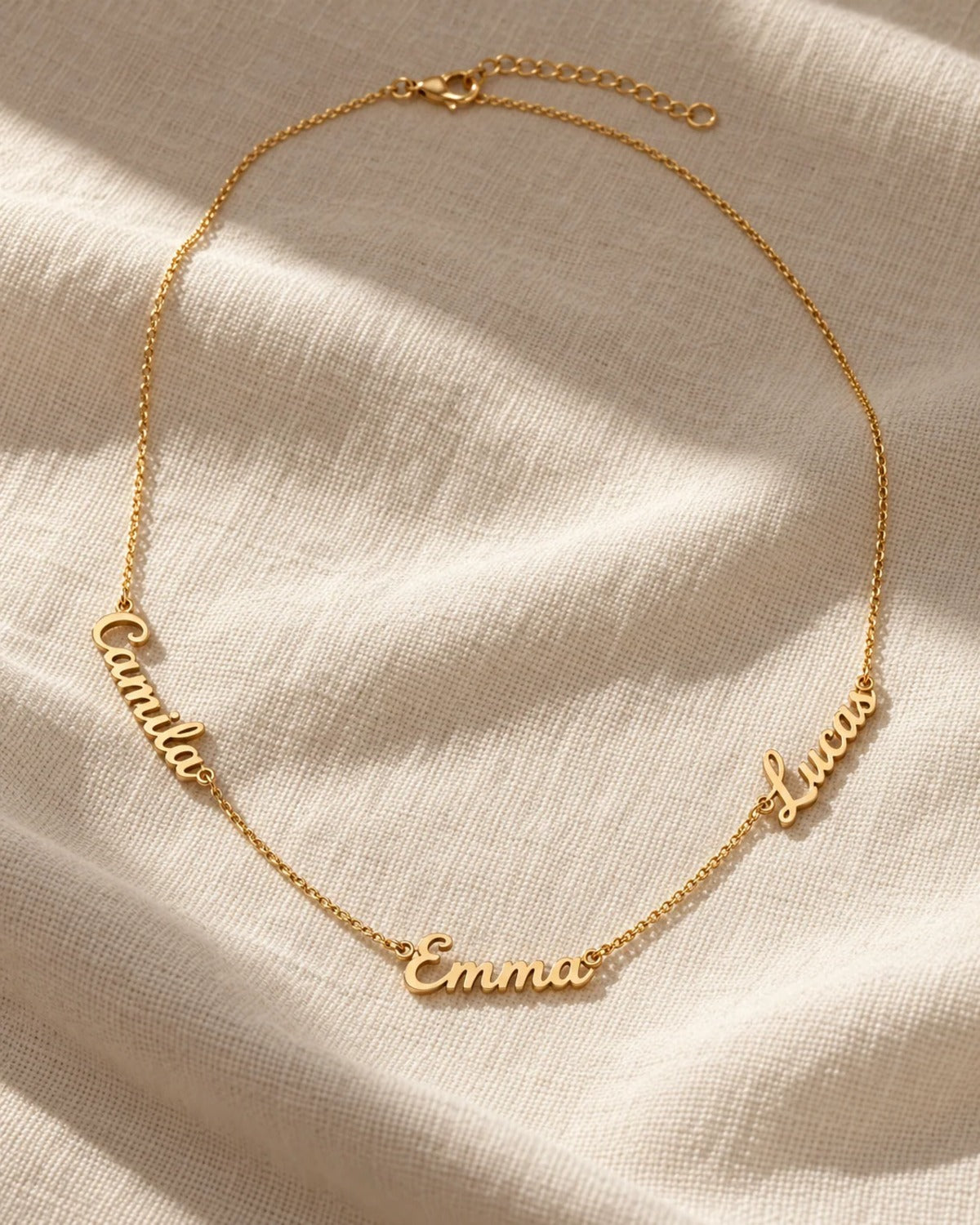 Gold name necklace with three cursive names, Camila, Emma and Lucas, on linen